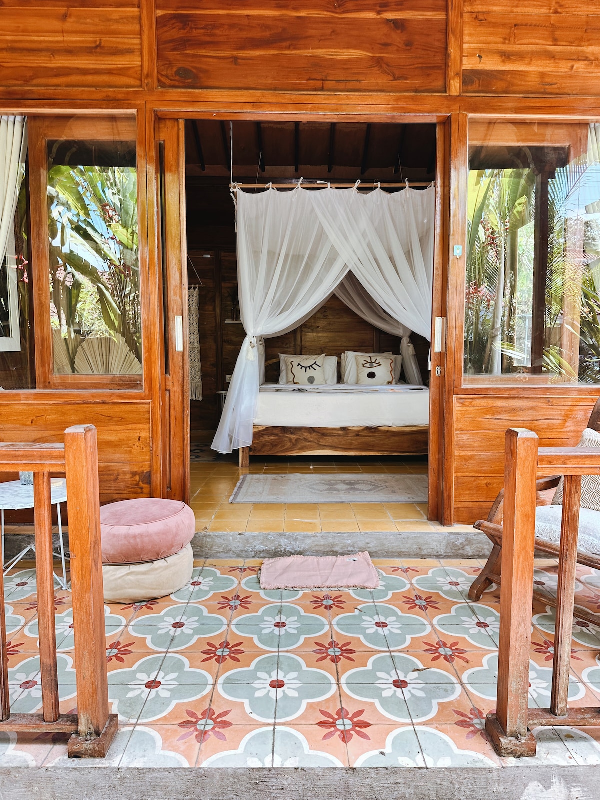The entrance to the bungalow is framed by wooden doors, leading to a cozy sleeping area with a kingsize bed draped in a white mosquito net. The outdoor patio features tiled flooring and simple seating, surrounded by lush greenery.