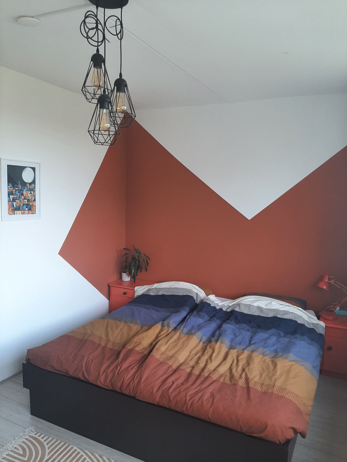 A bedroom is presented with a large bed covered in a multicolored striped blanket. The wall behind features a striking geometric design in earthy tones. Two bedside tables with lamps are positioned beside the bed, and a small potted plant adds a touch of greenery.