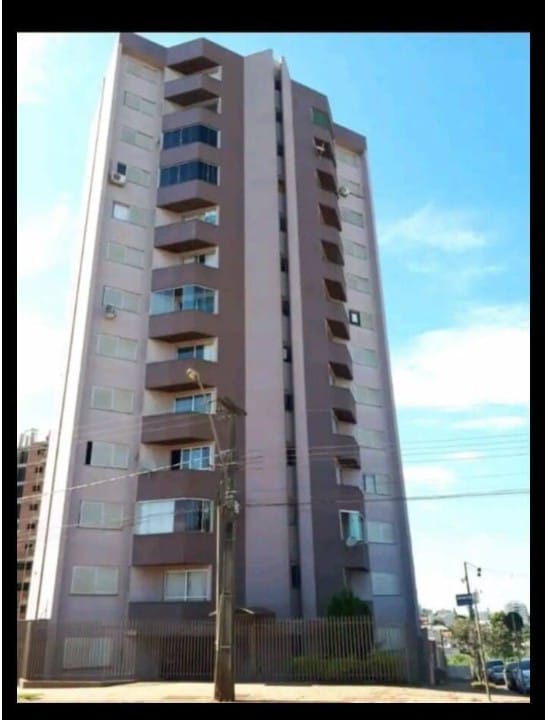 A modern five-story building is depicted, featuring a combination of flat and balcony windows. The façade displays a blend of beige and brown tones, with a mix of air conditioning units visible on the exterior. The surrounding area includes paved ground and some greenery.