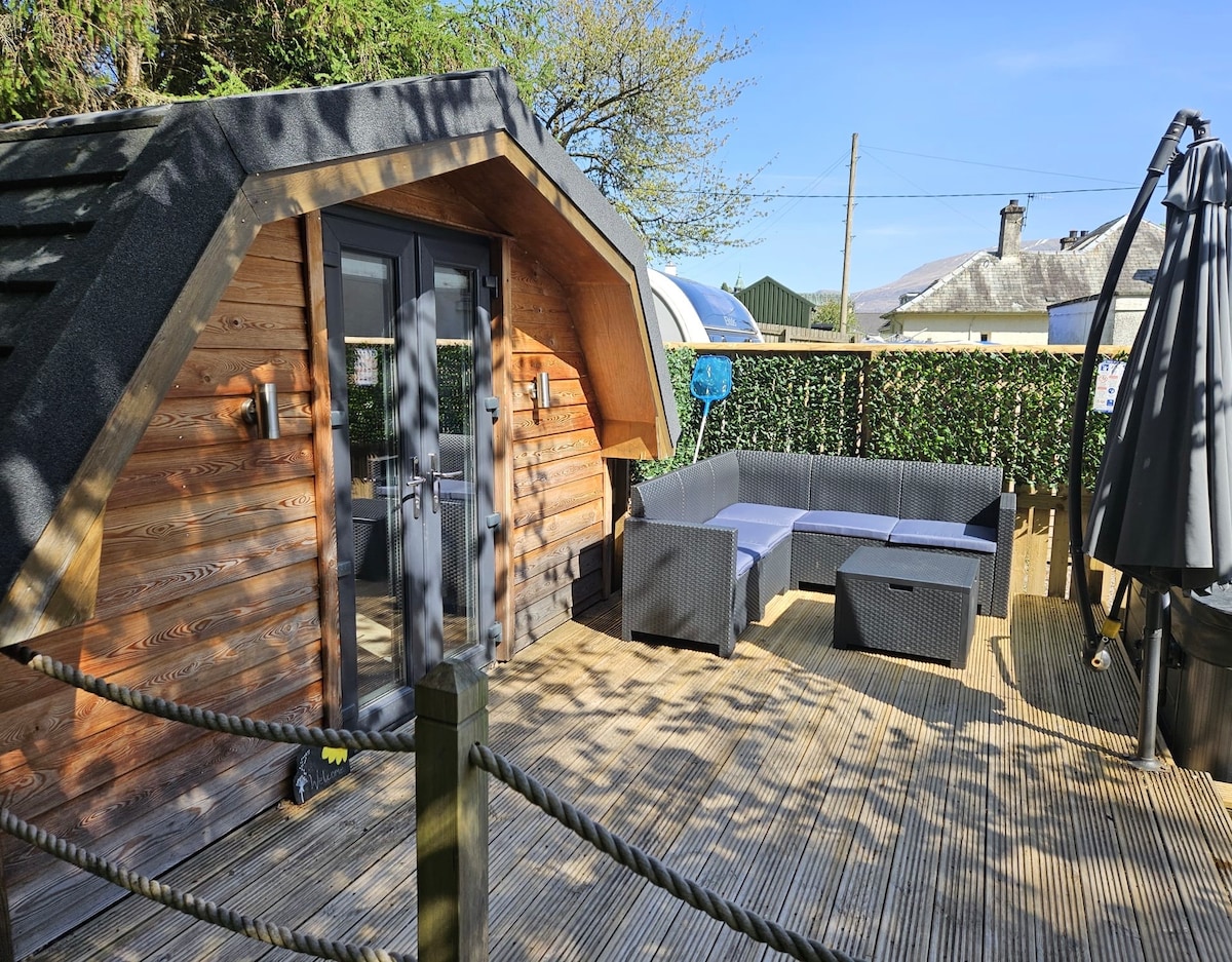 An outdoor seating area is featured, showcasing a sectional sofa with dark cushions set on wooden decking. The entrance displays double doors framed by wooden panels, and greenery is visible along the perimeter. A parasol provides shade in the corner, enhancing the inviting space.