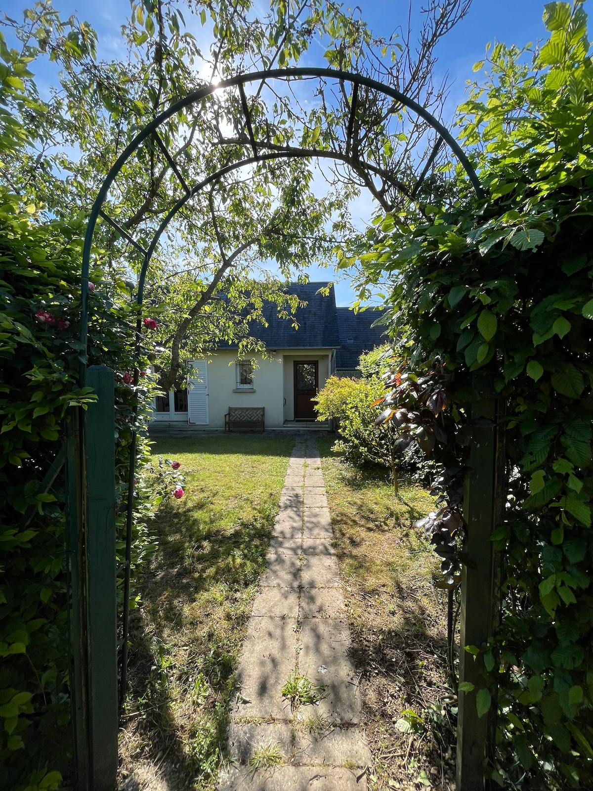 A garden entrance framed by greenery leads to a charming house. A stone path guides the way through the grass, flanked by blooming plants. The house is partially visible behind trees, with light filtering in from the clear blue sky.