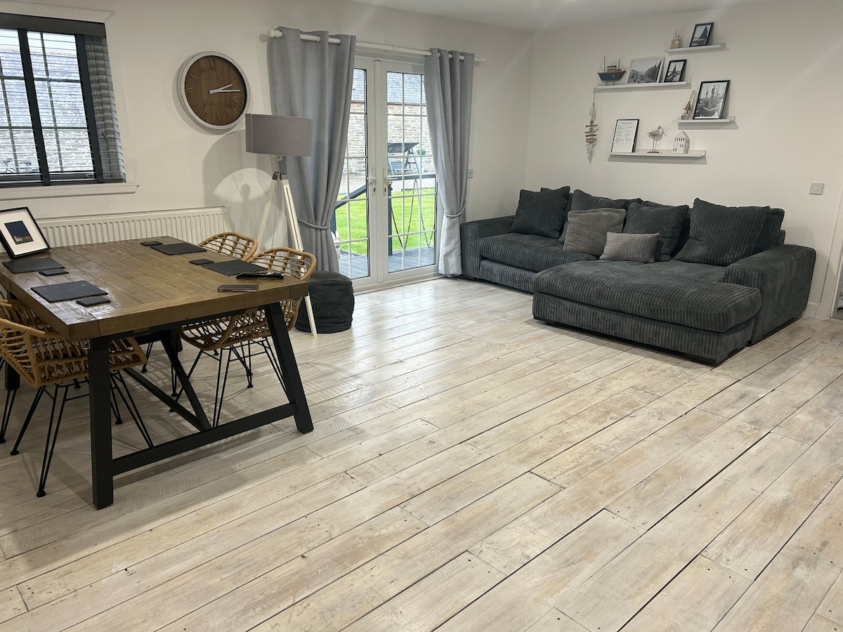 A comfortable living area features a spacious sectional sofa in a neutral tone and a wooden dining table surrounded by wicker chairs. Natural light enters through glass doors leading to the outdoors, while decorative shelves and a wall clock enhance the space's functionality.