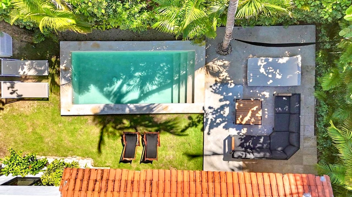 An aerial view captures a private pool accompanied by a shaded lounging area. Sunbeds are positioned near the pool, and a sleek seating arrangement is visible on a stone patio. Lush greenery surrounds the space, adding a refreshing touch to the outdoor setting.