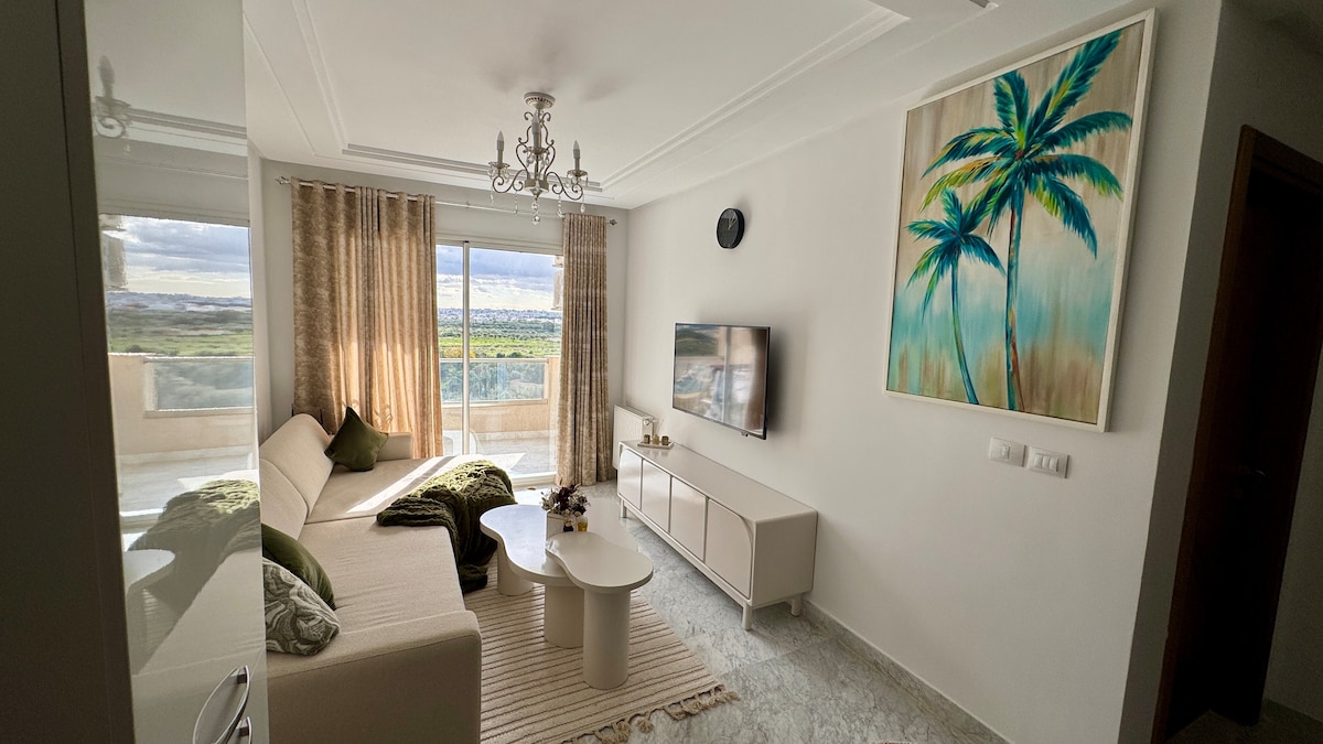 A modern living area is adorned with soft neutral-colored furnishings. Large windows allow natural light to fill the space, offering views of the surrounding landscape. A television is mounted on the wall, and a piece of tropical art adds a touch of color to the room.