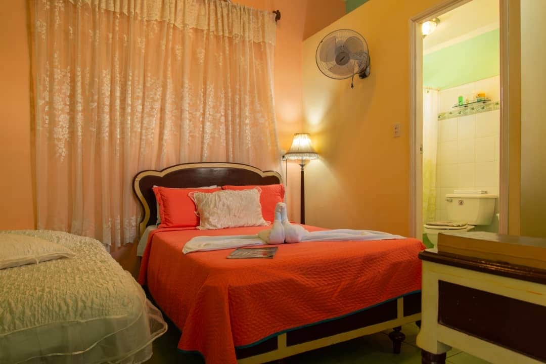 A vibrant bedroom is shown, featuring a spacious bed with coral-colored bedding. A smaller bed is placed beside it, accompanied by a lamp providing soft illumination. A bathroom door is visible in the background, highlighting convenience. Light filters through sheer curtains, adding warmth.