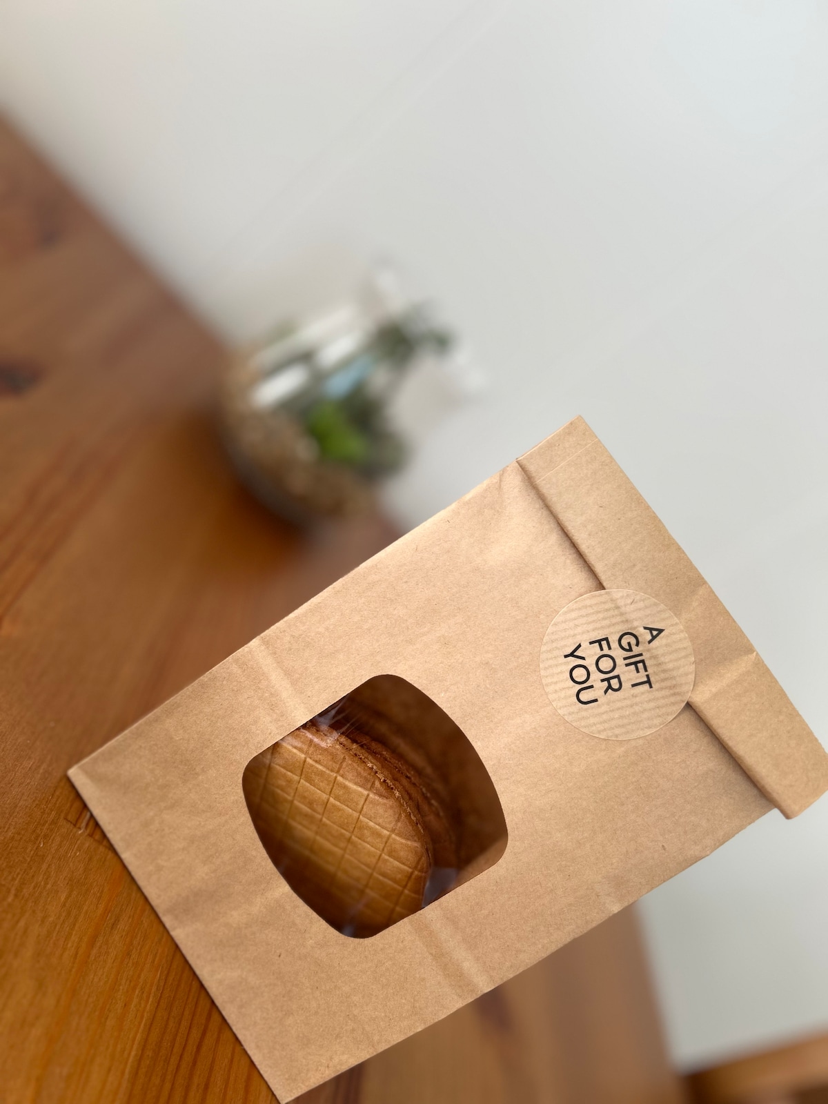 A brown paper bag is visible with a circular label reading 'A GIFT FOR YOU.' A round pastry is partially visible through the window of the bag, resting on a wooden surface. In the background, a small glass container with greenery is lightly blurred.