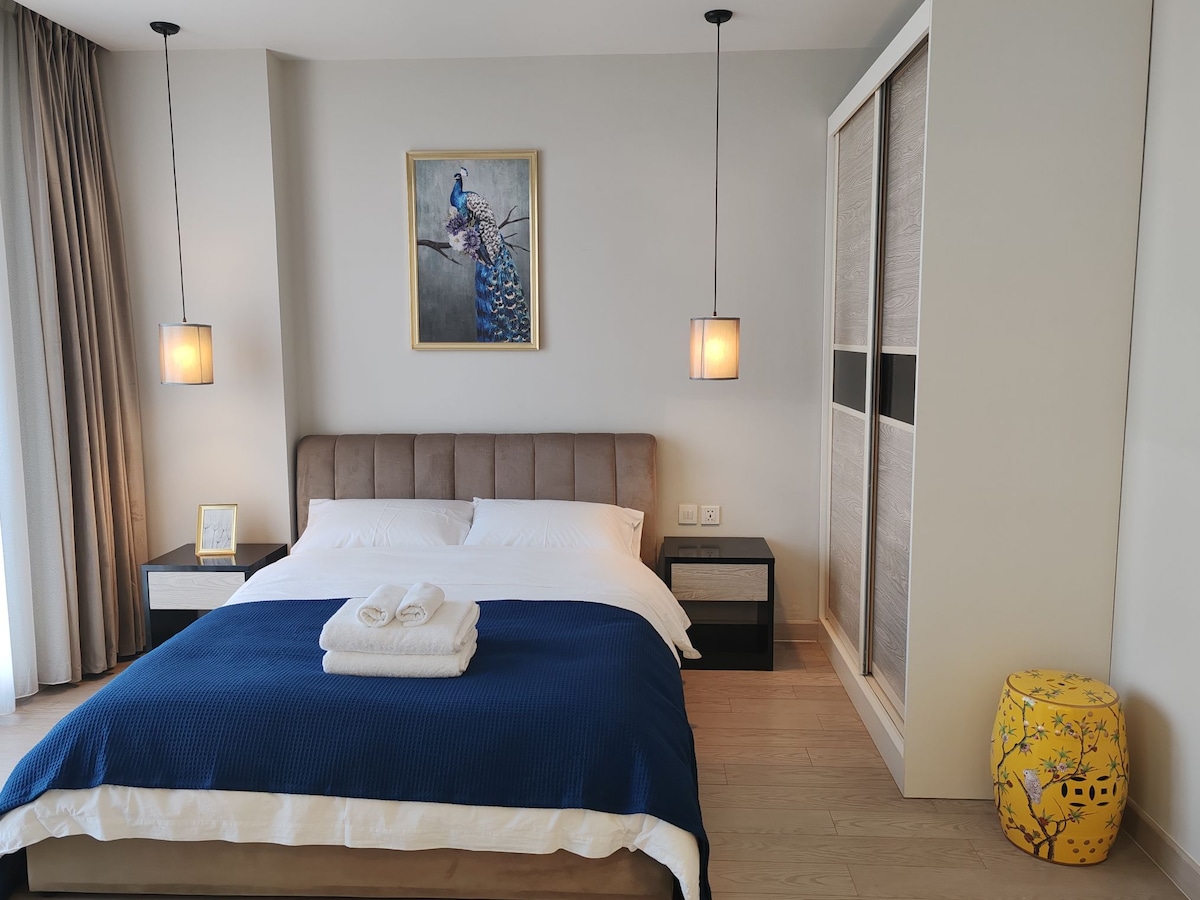 A spacious bedroom features a large bed with a textured blue blanket. Two bedside tables are positioned on either side, and a framed artwork of a peacock is displayed above the headboard. Warm lighting is provided by pendant lamps. A wardrobe is present in the corner.