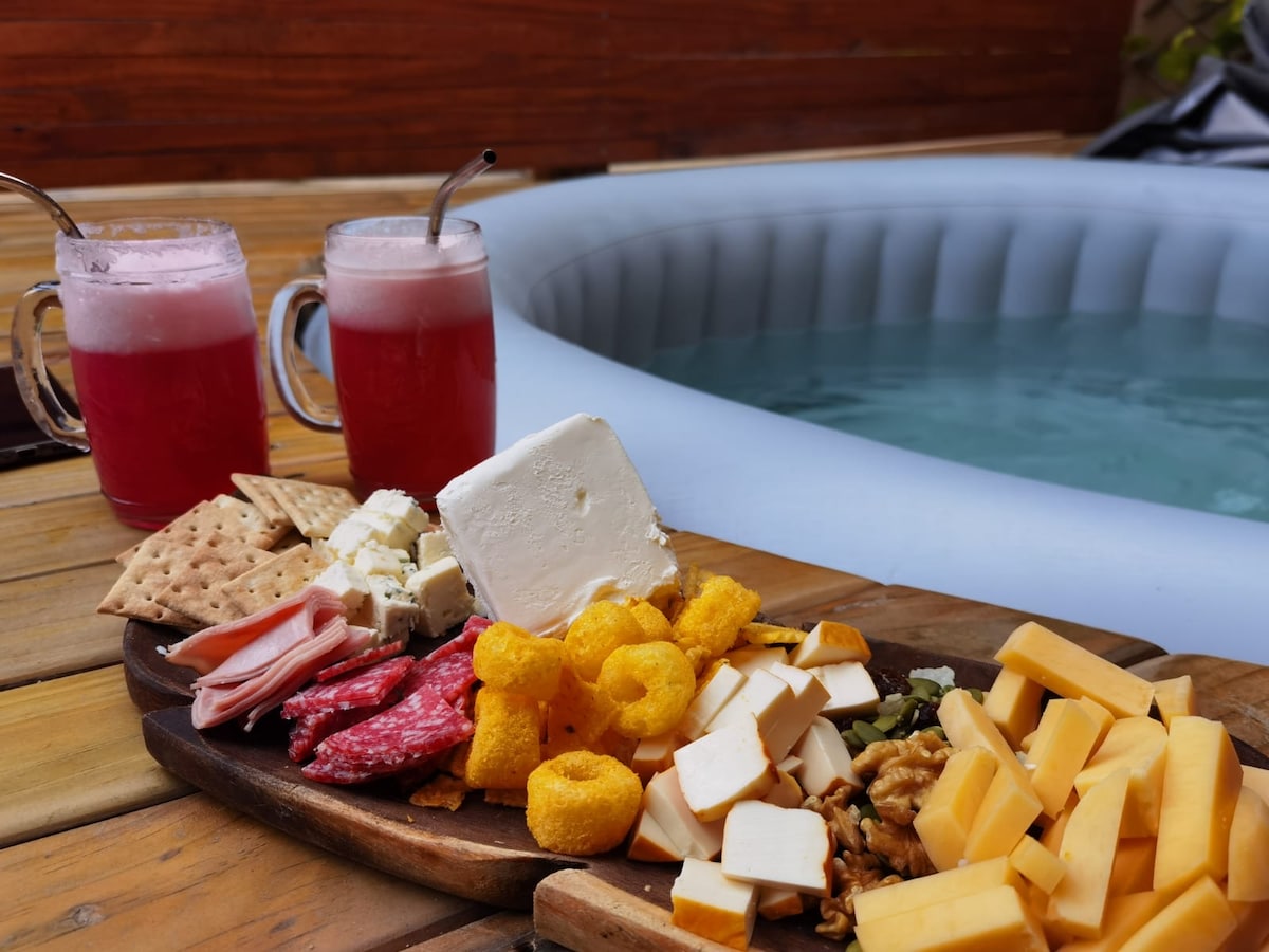A refreshing setup features two pink beverages in glass mugs beside an assortment of cheeses, cured meats, and snacks arranged on a wooden platter. The background showcases a jacuzzi, inviting relaxation and enjoyment.