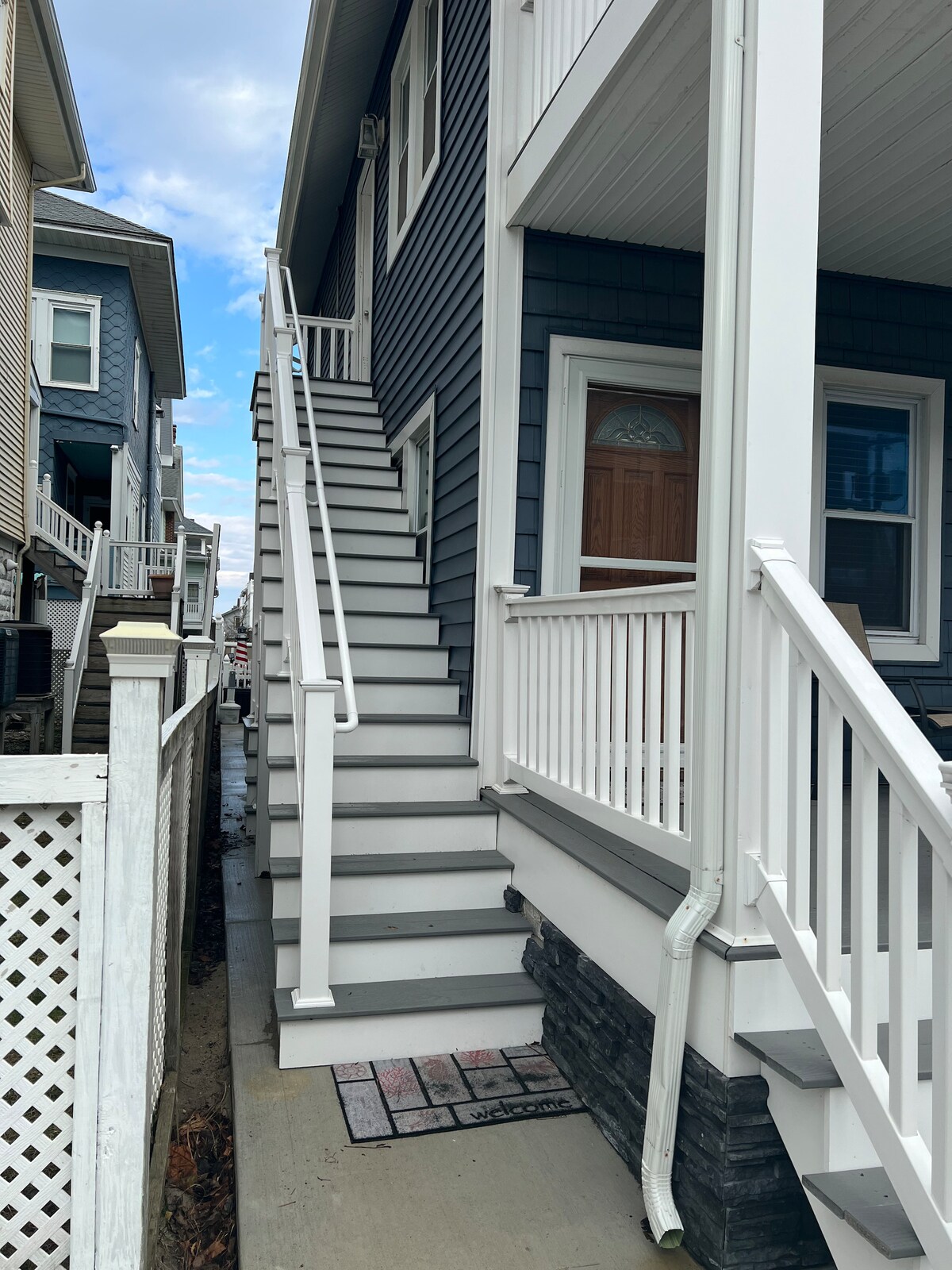 A set of white stairs leads up to a condo entrance, flanked by a grey-shingled building. The clean and well-maintained exterior is complemented by a white railing and porch area. Surrounding structures exhibit similar coastal architecture.
