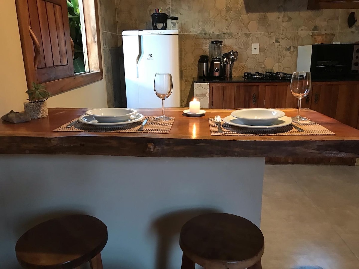 A wooden counter is set with two plates and wine glasses, accompanied by a lit candle. In the background, a well-equipped kitchen features a refrigerator and cooking appliances, highlighting the functionality of the space.