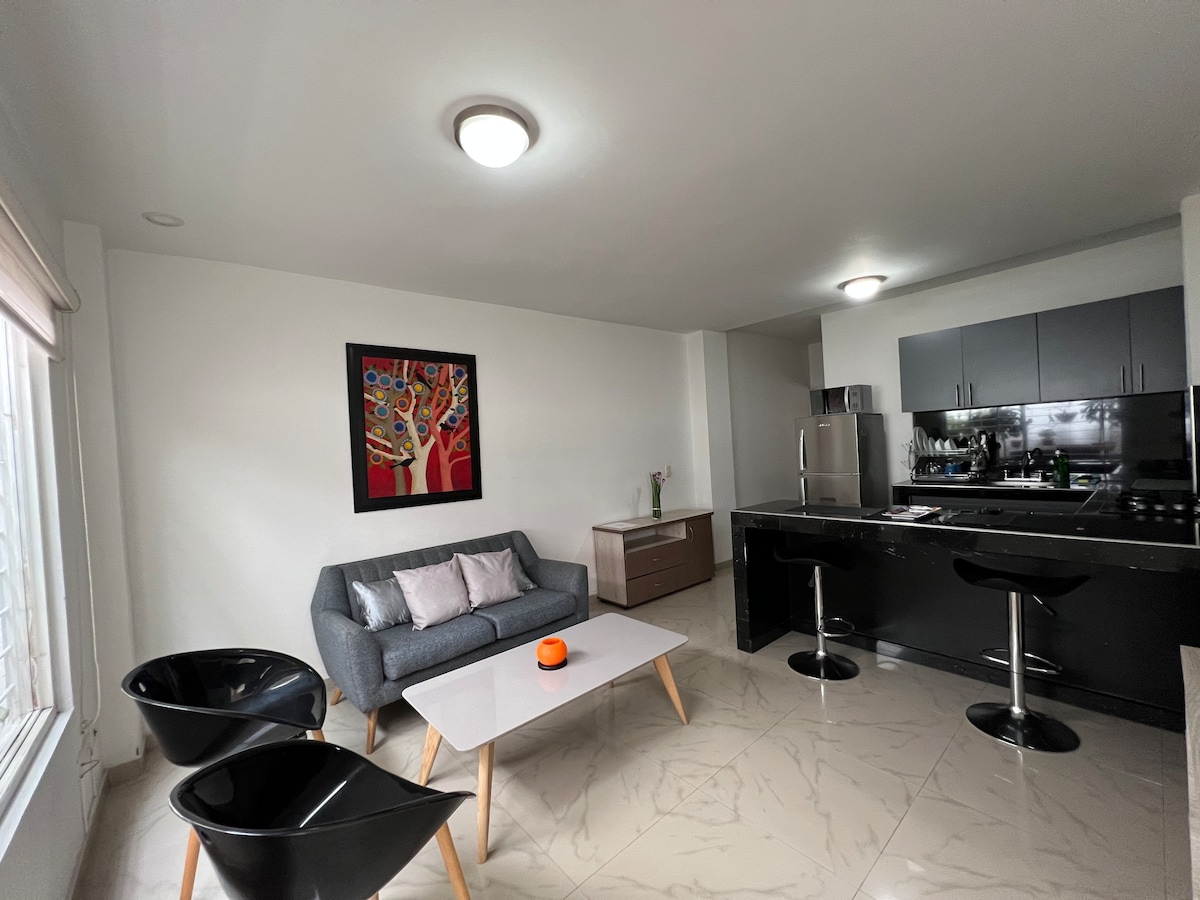 A modern living area features a grey sofa and two black armchairs. A coffee table sits centered, highlighted by an orange decoration. An abstract painting adorns the white wall, and the kitchen area is visible, showcasing sleek cabinetry and stainless-steel appliances.