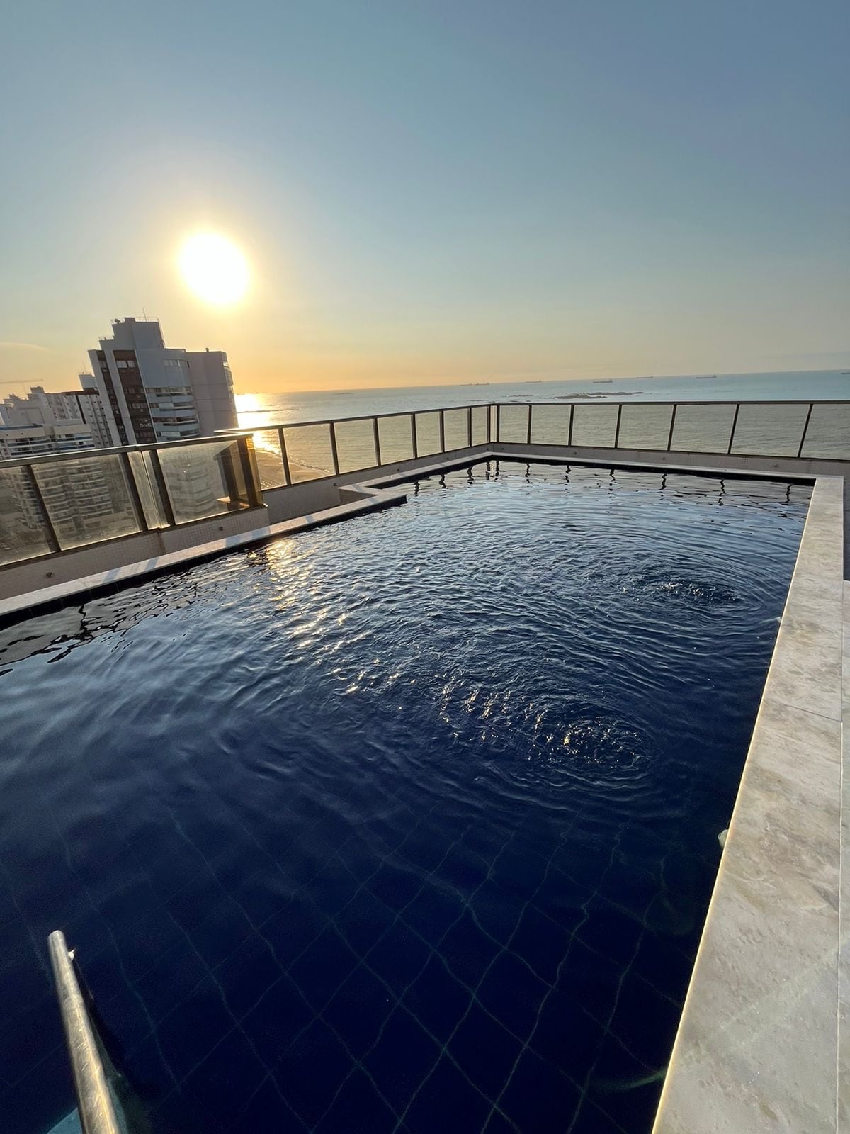 A rooftop pool glistens under the setting sun, surrounded by a glass railing that provides unobstructed views of the horizon. The calm water reflects warm hues from the sky, creating a serene atmosphere. Nearby buildings are faintly visible in the background.
