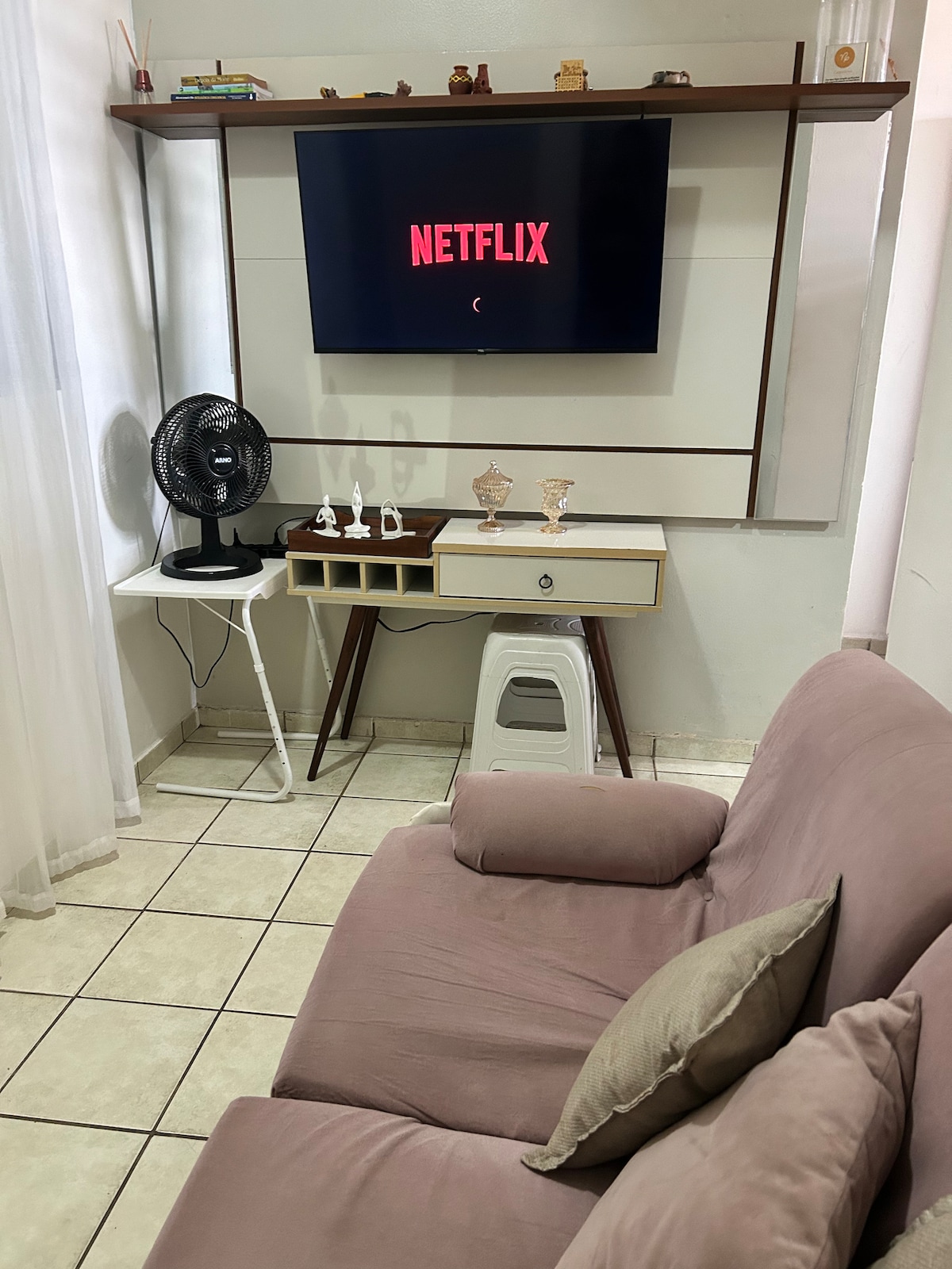 A cozy living area features a soft pink sofa paired with a small white table. A fan is positioned nearby, and a television mounted on the wall displays the Netflix logo. Light curtains allow natural light to filter in, illuminating the space.