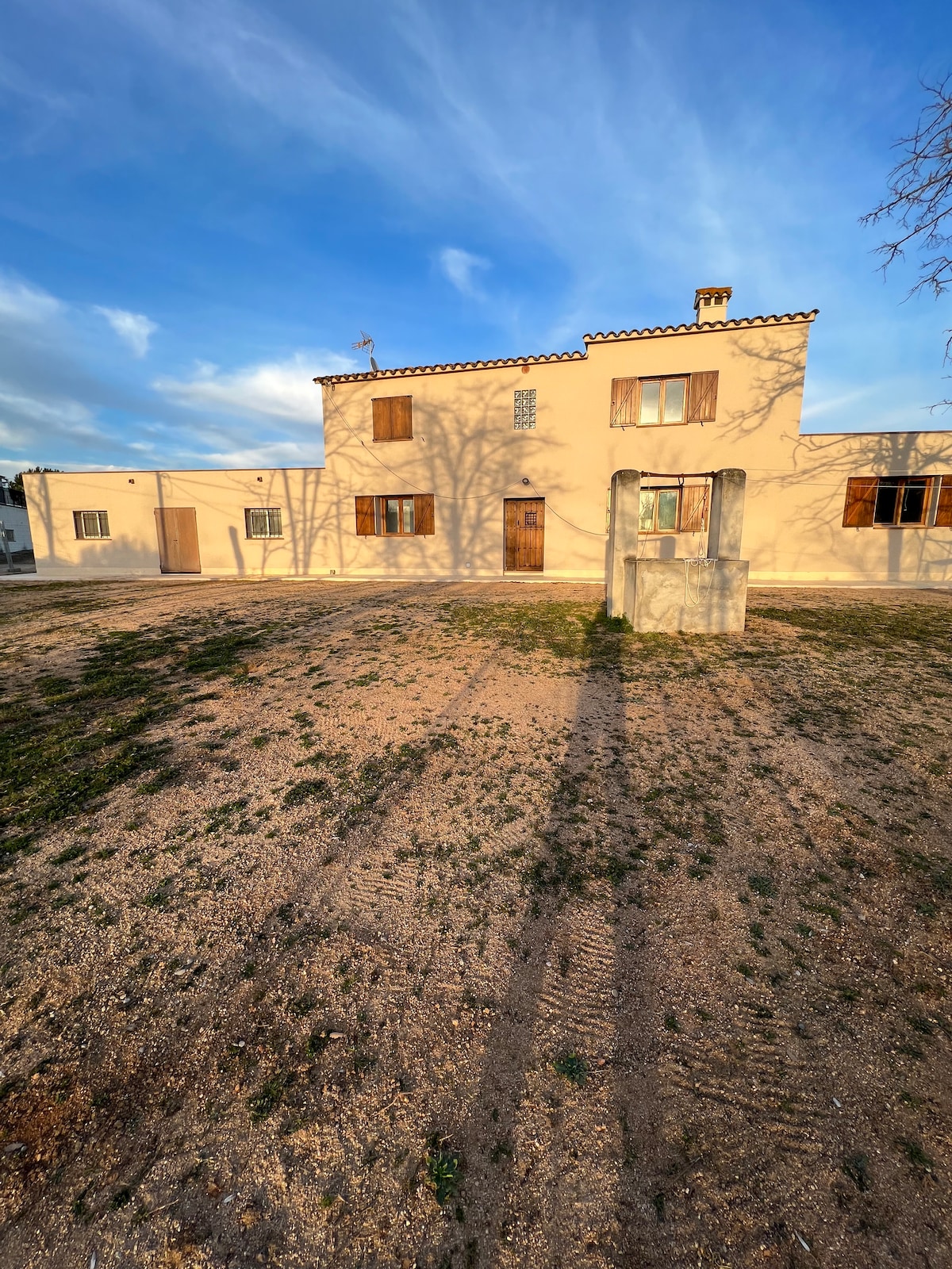 A spacious rural home is shown under an open sky, with a light-colored facade and multiple windows. A stone well is visible in the foreground, with shadows cast by the house and surrounding trees enhancing the serene atmosphere of the natural setting.