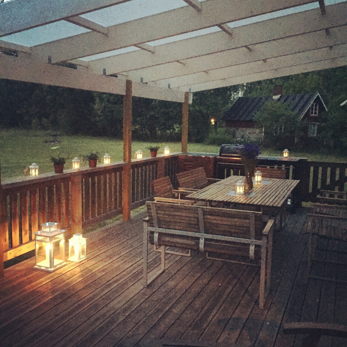 A spacious wooden deck is shown, featuring a large table surrounded by several chairs. Soft lighting from lanterns casts a warm glow, creating an inviting atmosphere. Potted plants are arranged along the railing, complemented by a barbecue grill in the background.