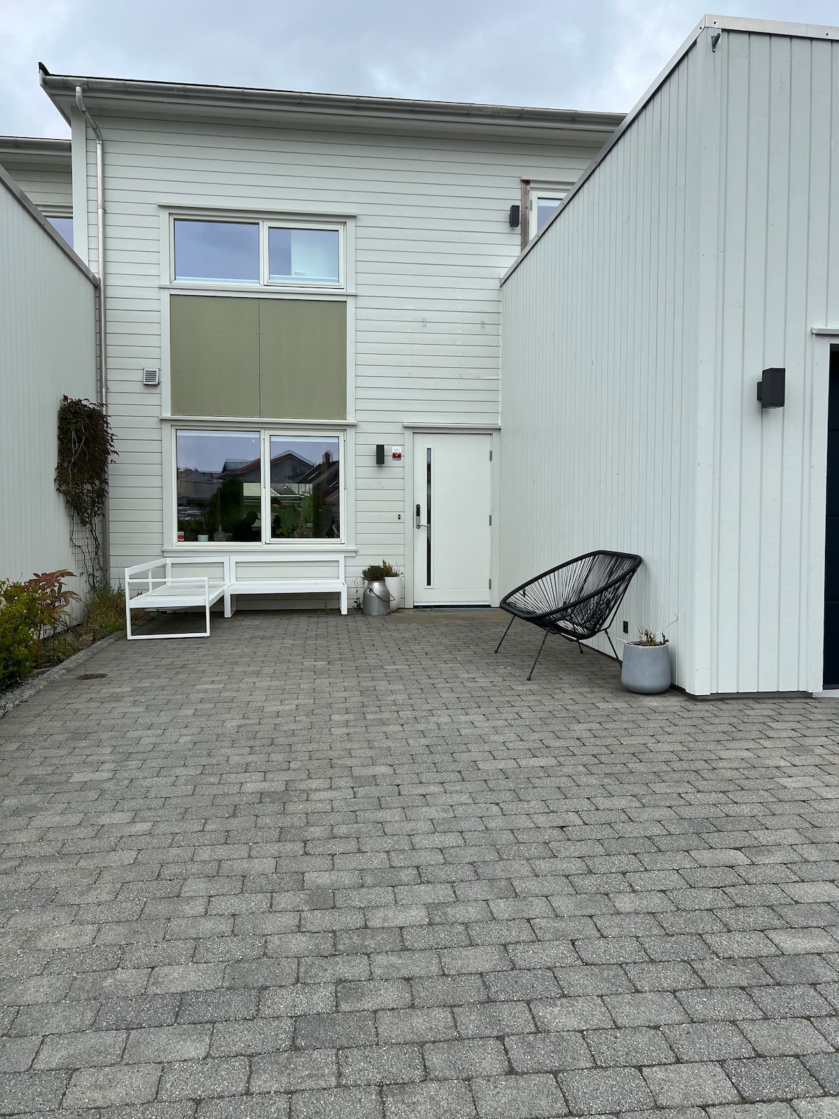 An exterior view of the house shows a paved area leading to a bright entrance. A large window allows natural light inside, while a white outdoor bench and a black chair are positioned on the paved surface for seating. Greenery is present beside the entrance.