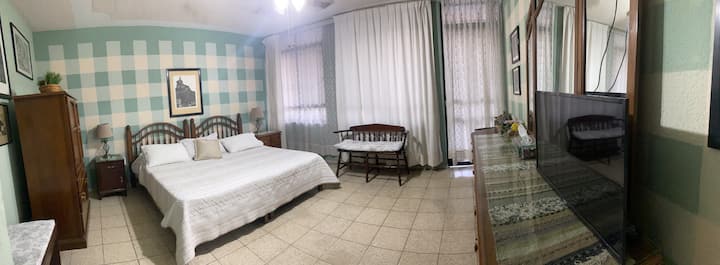 ห้องนอนขนาดใหญ่พร้อมเตียงคิงไซส์ การตกแต่งในโทนสีเขียวและไม้ หน้าต่างบานใหญ่ที่ให้แสงธรรมชาติ และระเบียงพร้อมเก้าอี้และโต๊ะ มีทีวี พัดลมติดเพดาน และพื้นที่สบายสำหรับการพักผ่อนในบรรยากาศที่อบอุ่นและเงียบสงบ