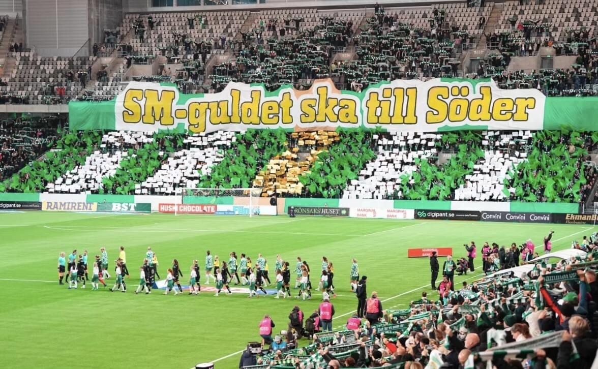 A vibrant crowd fills the stadium, showcasing a large display of colorful banners and coordinated fan movements. Green, yellow, and white colors dominate the scene, creating a spirited atmosphere as players enter the field, setting the stage for an exciting football match.
