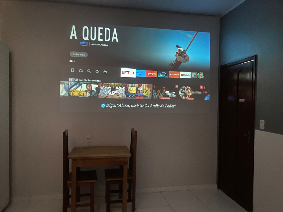 A wall features a large projection displaying a streaming service interface, showcasing various available movies and shows. A small table with two chairs is positioned beneath the projection, creating an inviting area for entertainment.