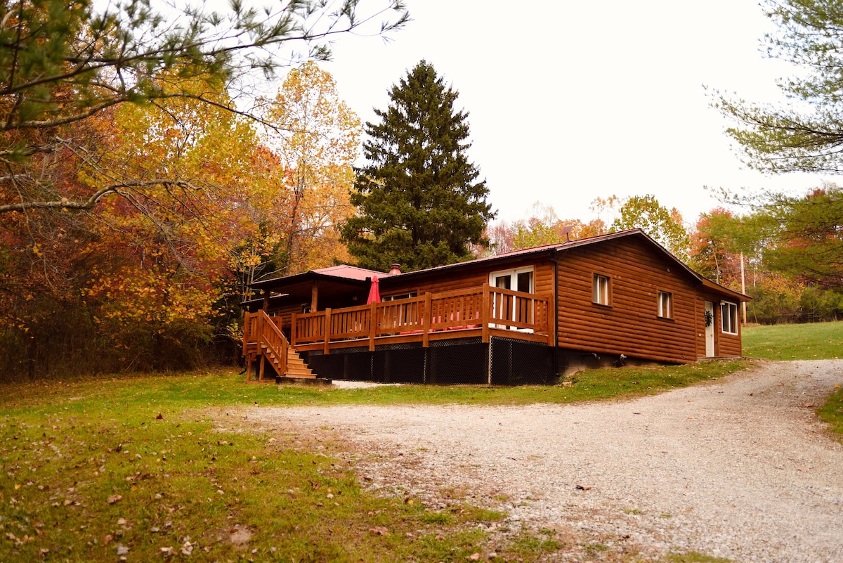 A wooden cabin is nestled in a natural setting, featuring a spacious porch with a railing. The entrance is accessible via a set of stairs. Surrounding trees display autumn colors, and a gravel driveway leads up to the cabin.