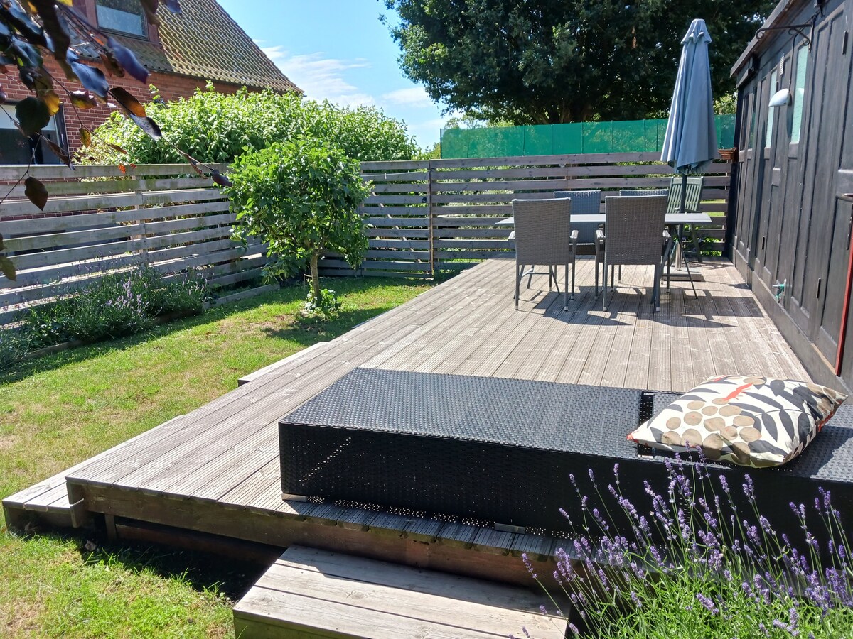 An outdoor terrace is presented with wooden decking and a spacious seating area featuring a table and chairs. A sun lounger is positioned nearby, complemented by a decorative pillow. A green lawn surrounds the setup, with lavender plants adding subtle color. A sunshade offers additional comfort.