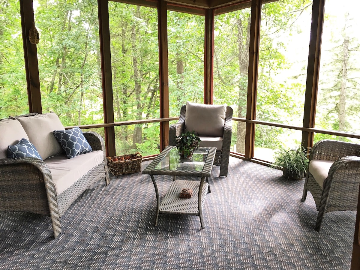 A screened-in porch is surrounded by lush greenery, featuring comfortable seating with throw pillows. A glass coffee table rests at the center, while a decorative basket and a small potted plant add natural accents to the space.