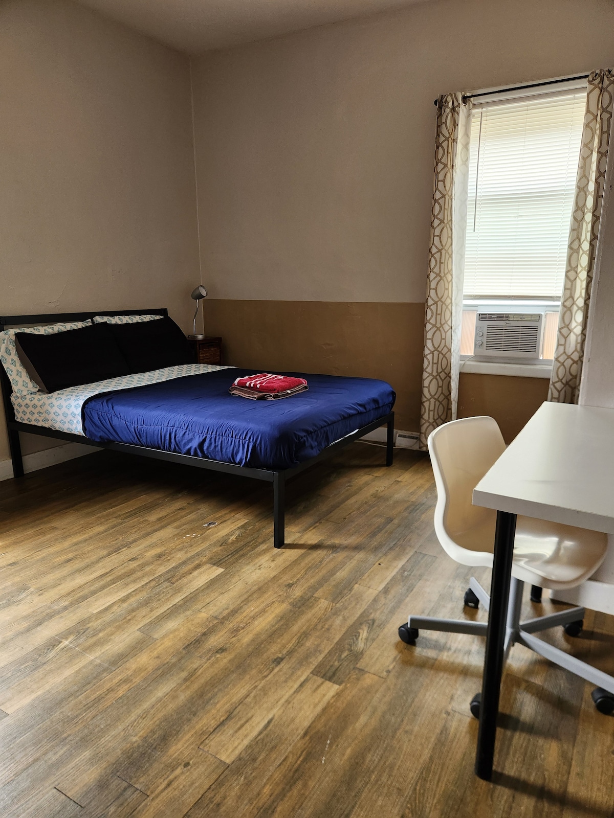 A private bedroom is equipped with a metal frame bed, featuring a navy blue fitted sheet and a patterned headboard. A small lamp is positioned on a bedside table. A desk with a white chair is located near a window, which is adorned with patterned curtains.