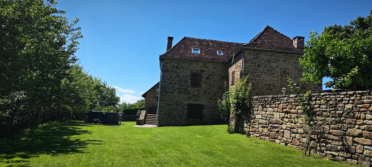 The exterior of a charming stone house is depicted, featuring a well-maintained grassy area. Trees provide natural shade, while the structure showcases multiple windows and a distinctive roof. A stone wall borders the property, enhancing the sense of privacy and tranquility.