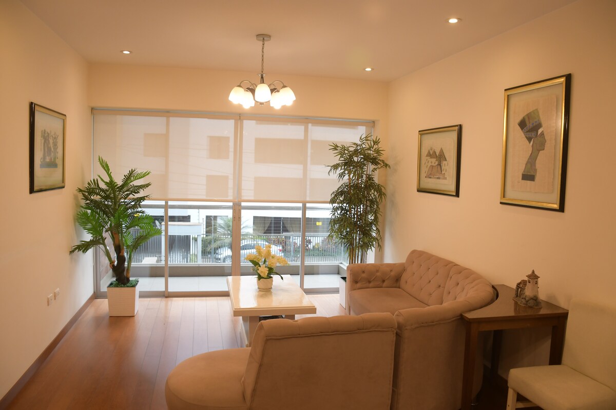 A bright living area features large windows that offer a view of the surroundings. A decorative plant stands beside a stylish beige sofa and armchair. A small table is centered in the space, accented by a floral arrangement. Framed artwork adorns the walls.