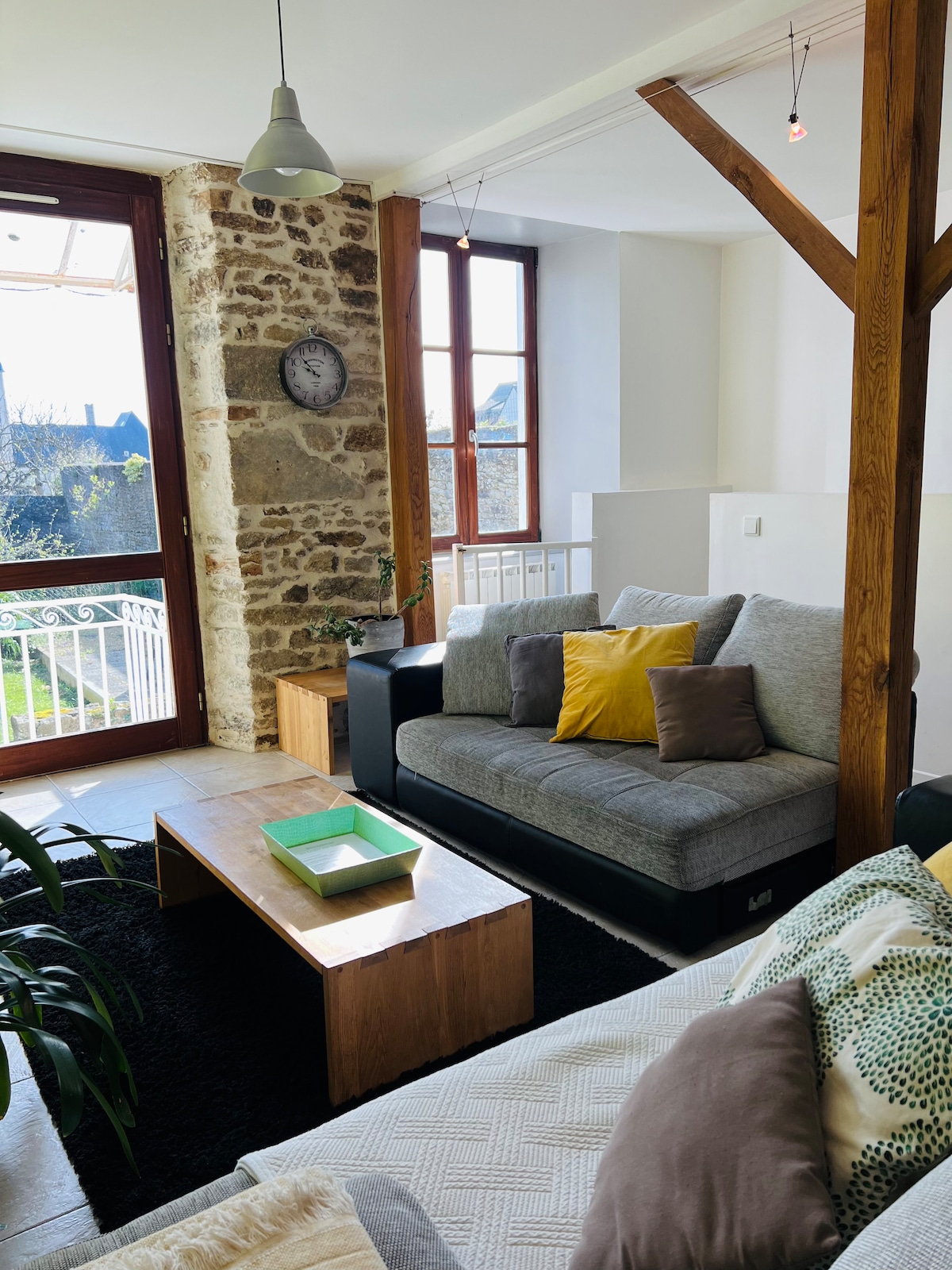 A cozy living space is highlighted by a gray sofa adorned with colorful cushions. Natural light filters through large windows, casting soft shadows on the wooden coffee table and carpet. Exposed stone walls add a touch of rustic charm to the overall design.