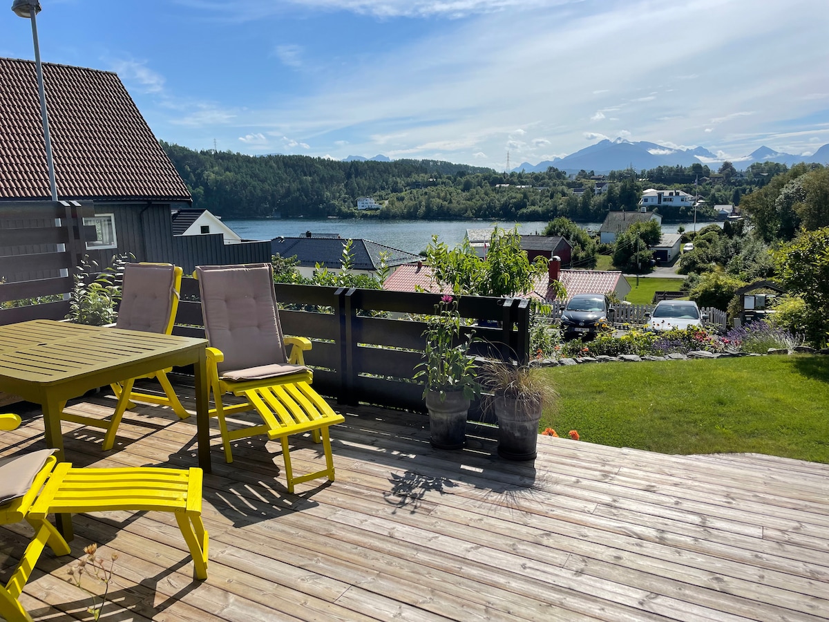 A sunlit terrace is furnished with yellow lounge chairs and a table, surrounded by potted plants. The view showcases a serene landscape of greenery and water, with distant mountains forming a picturesque backdrop. The wooden deck enhances the outdoor experience.