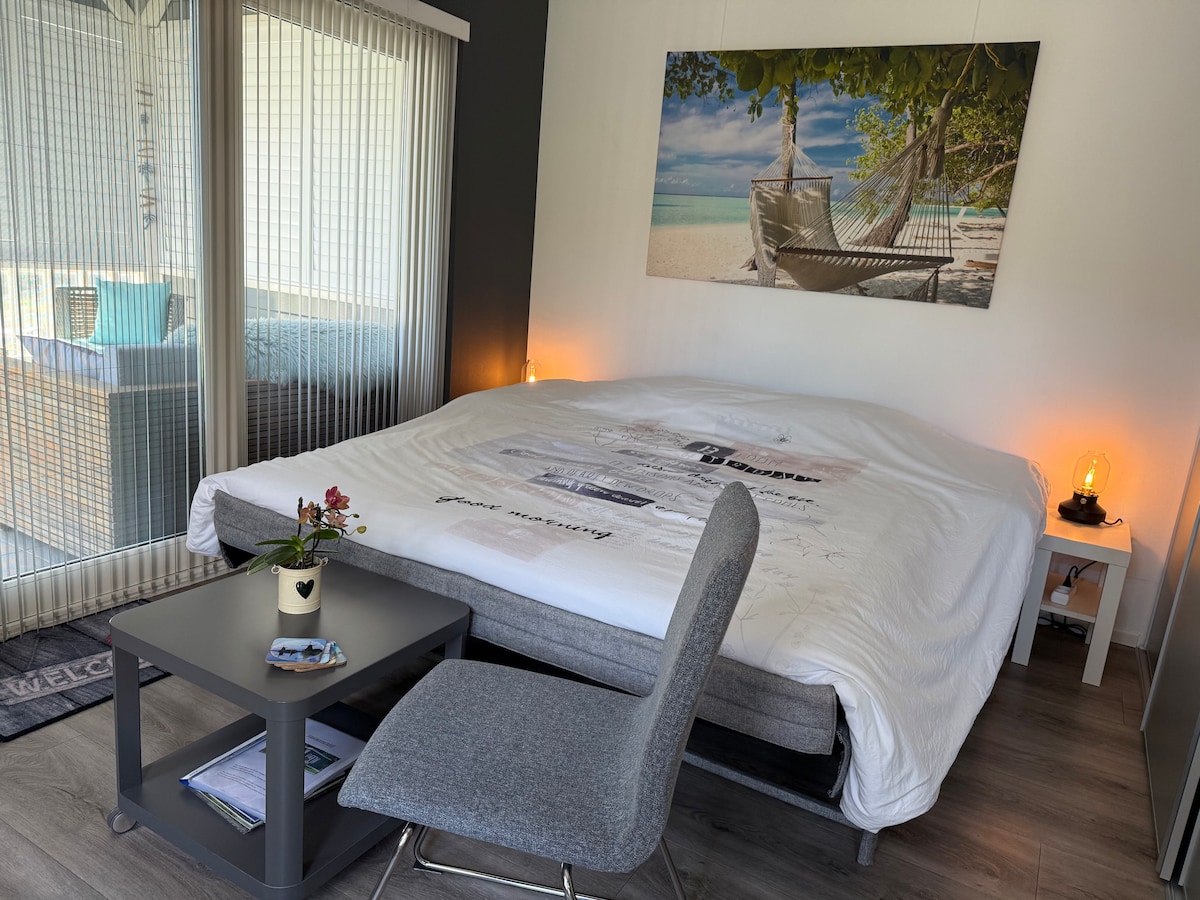 A modern sleeping area features a spacious bed with light bedding, complemented by bedside lamps. A small table sits near a grey chair, with a wall-mounted photograph depicting a serene beach scene. Natural light enters through sheer curtains, creating a calm environment.
