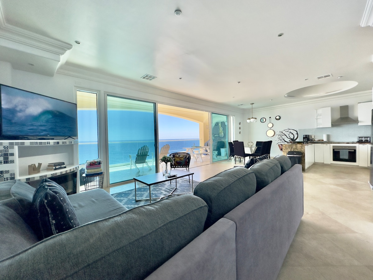 The open living space features a large grey sofa facing a modern television. Floor-to-ceiling windows provide ocean views, with light streaming into the area. The adjoining dining space includes a dark table surrounded by comfortable chairs, offering an inviting area for meals.