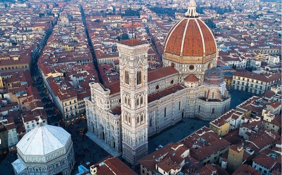 An aerial view captures the iconic landmarks of Florence, showcasing the majestic dome of the Cathedral of Santa Maria del Fiore and the adjacent Giotto's Campanile. Red-tiled roofs span the city, creating a harmonious blend of historical architecture against a backdrop of fading daylight.