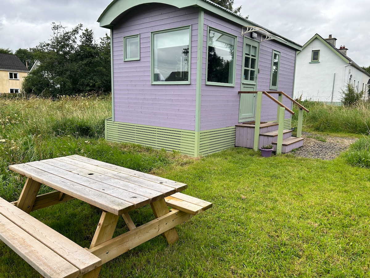 The shepherd's hut is shown in soft purple tones, surrounded by lush green grass. A wooden picnic table is positioned in the foreground, offering a quaint outdoor dining area. The hut features large windows, providing ample natural light and views of the countryside.