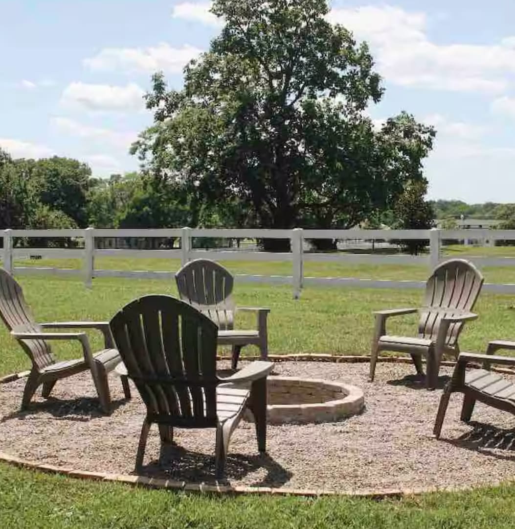 A fire pit surrounded by a gravel circle is set in a grassy area, featuring several wooden chairs arranged for seating. A large tree and white fencing are visible in the background, contributing to a serene outdoor setting.