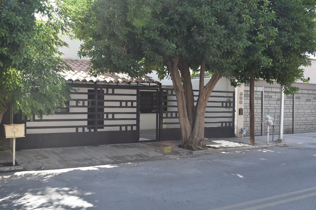 The exterior of the house features a modern design with a combination of bold black and white panels, providing a striking contrast against the lush greenery of nearby trees. A gated entrance indicates a secure environment, complemented by a visible driveway.