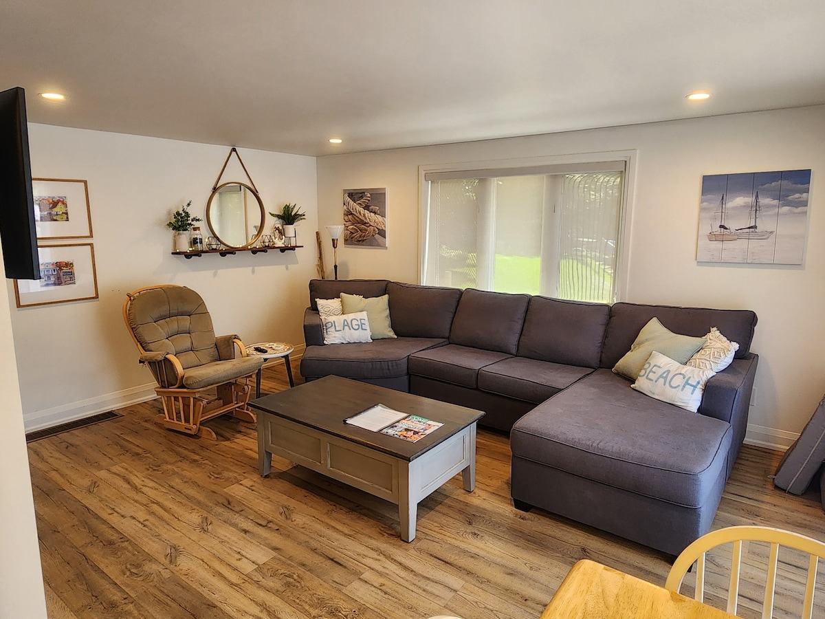 The living room features a spacious gray sectional sofa, complemented by a comfortable rocking chair. A large coffee table occupies the center, and natural light fills the space through large windows. Beach-themed decor enhances the relaxed atmosphere.