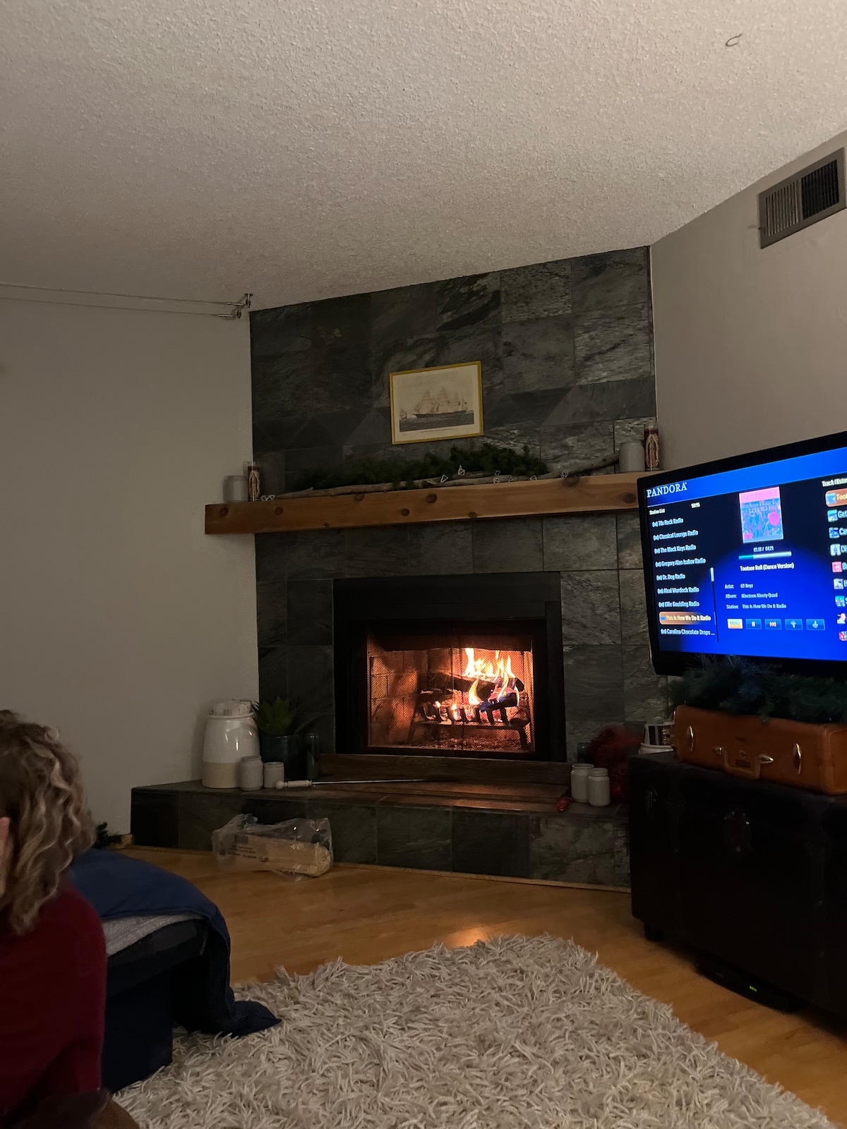 A cozy living area is depicted with a stone fireplace emitting warm flames. A wooden shelf is mounted above the fireplace, adorned with decorative items. A spacious area rug covers the wooden floor, and a television screen displays various content. Soft lighting enhances the room's ambiance.