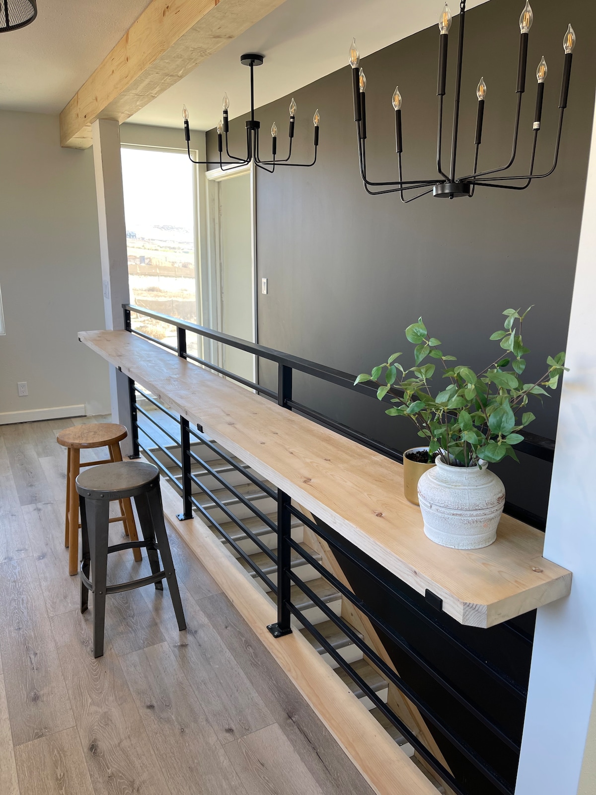 A stylish wooden counter is paired with two simple stools, enhancing the open space. A modern chandelier with several bulbs hangs overhead, while a potted plant adds a touch of greenery. Large windows provide natural light and a view of the surrounding landscape.