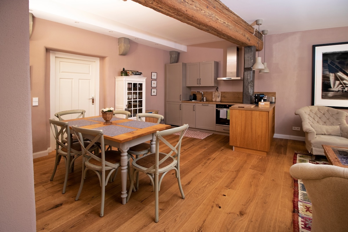 A spacious dining area features a rustic table surrounded by six light-hued chairs. Modern kitchen appliances are seamlessly integrated into the design. Exposed wooden beams and soft wall colors enhance the warm ambiance of this inviting living space.