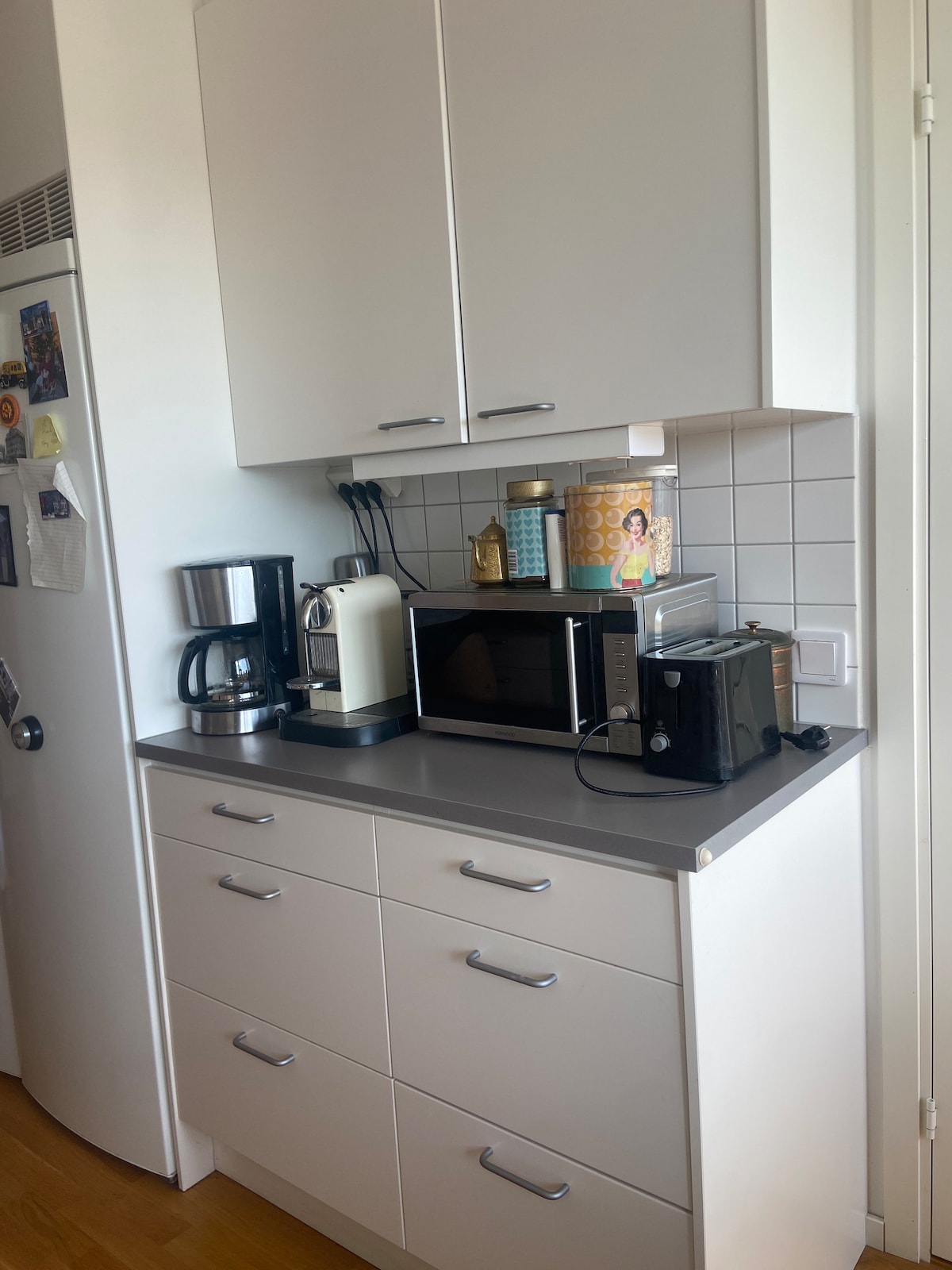 A compact kitchen area features modern appliances including a coffee maker, microwave, and toaster. Storage cabinets in white complement the light grey countertop. A refrigerator is visible on the left, and a decorative coffee container adds a touch of functionality.