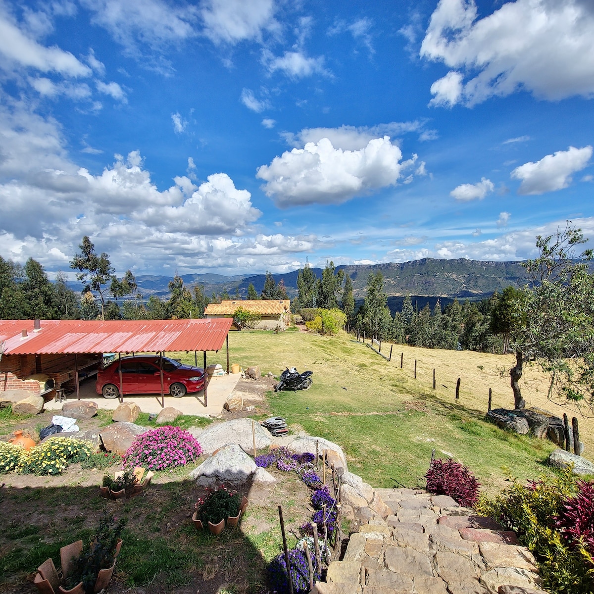 A panoramic view showcases a well-maintained outdoor space with a red-roofed structure providing shade. A car is parked nearby, surrounded by lush green grass and an array of colorful flowers. The distant mountains and expansive sky add a scenic backdrop.