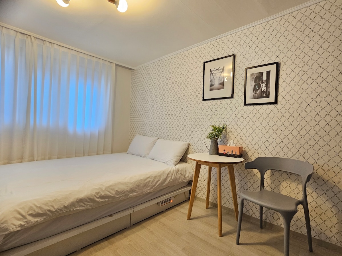 A clean and cozy bedroom features a spacious bed with white linens and two pillows. A small round table is accompanied by a grey chair, and framed black-and-white photos adorn the walls. Soft natural light enters through sheer curtains, creating a bright atmosphere.