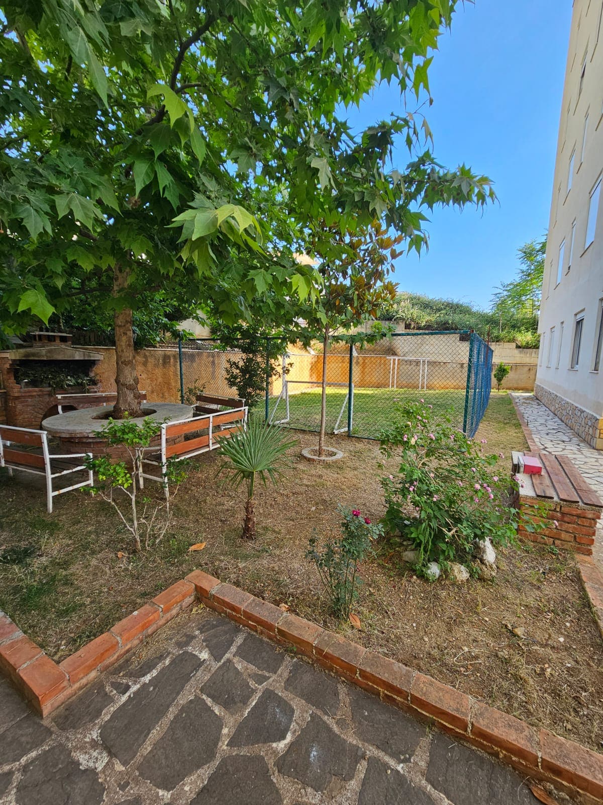 A serene garden area is depicted, featuring a large tree providing shade. A small seating space surrounded by wooden benches and greenery is visible. A fenced area is shown in the background with grass and various plants, contributing to the peaceful atmosphere.