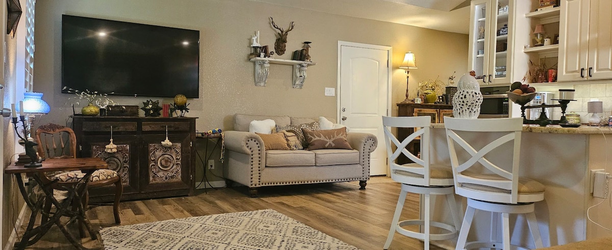 The living area features a beige sofa adorned with decorative pillows, positioned near a mounted television. A wooden table and chairs provide seating near the kitchen. Natural light fills the space through window coverings, enhancing the warm tones of the room.