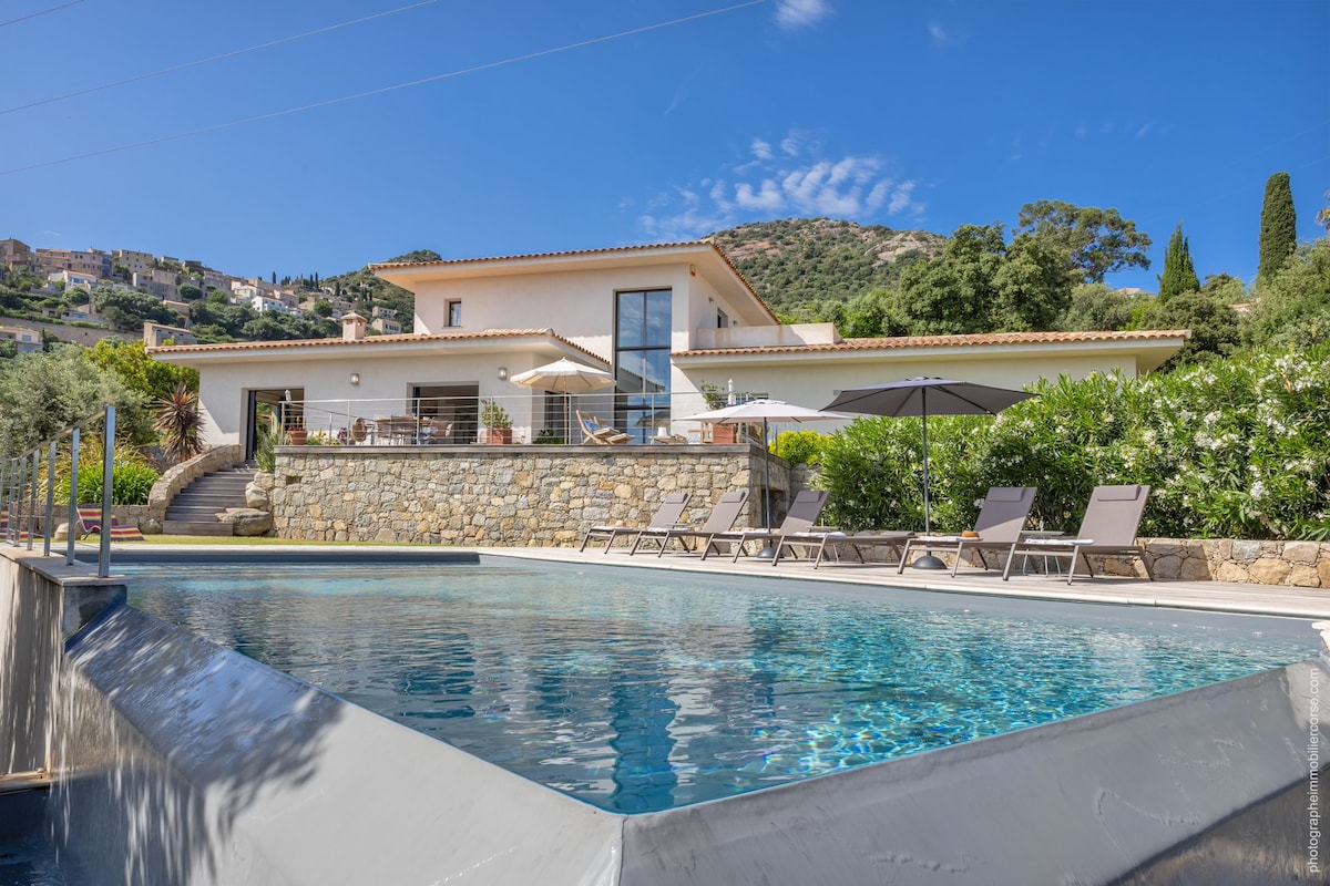 A modern architectural house is set against a backdrop of greenery, featuring a striking infinity pool. The pool reflects the clear blue sky and is surrounded by lounge chairs. A spacious terrace with shaded seating areas enhances the outdoor experience.