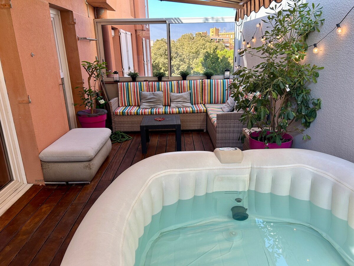 A terrace area features a jacuzzi surrounded by a stylish seating arrangement. Colorful striped cushions adorn the seating, complemented by potted plants. Warm string lights are positioned overhead, creating a cozy ambiance. The wooden deck provides a natural look, enhancing the outdoor space.