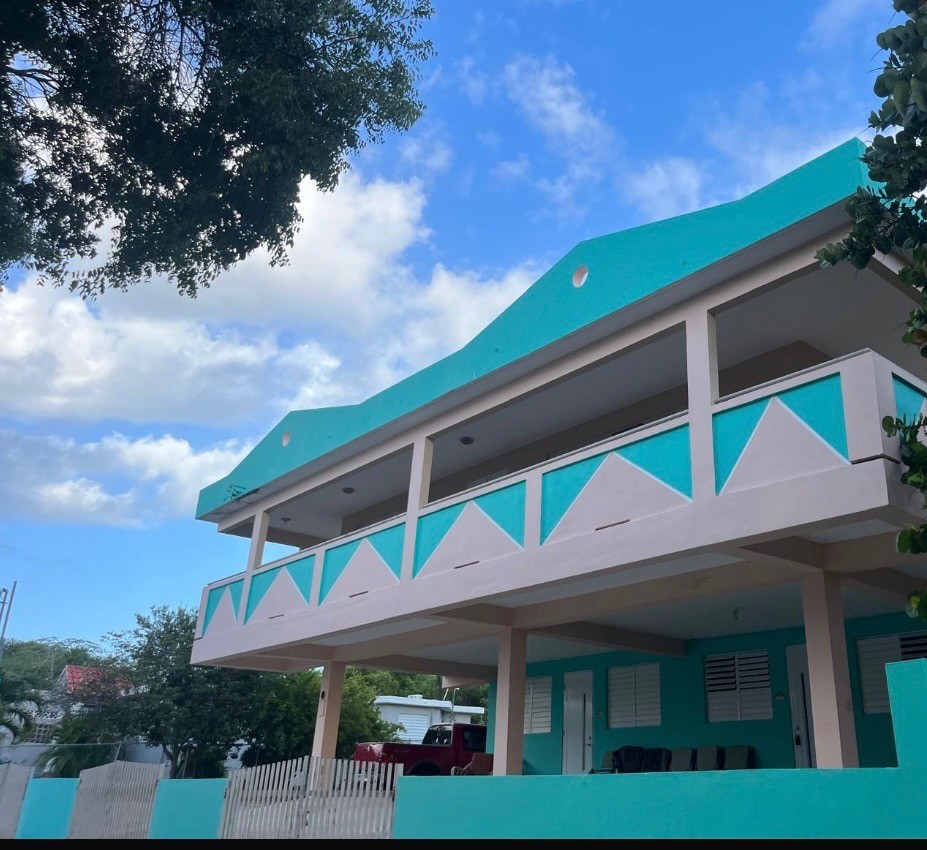 A two-story building features bright turquoise walls with white geometric accents. The balcony overlooks the ground floor, which is shaded by trees. A white picket fence can be seen in front of the property, adding charm to the exterior.