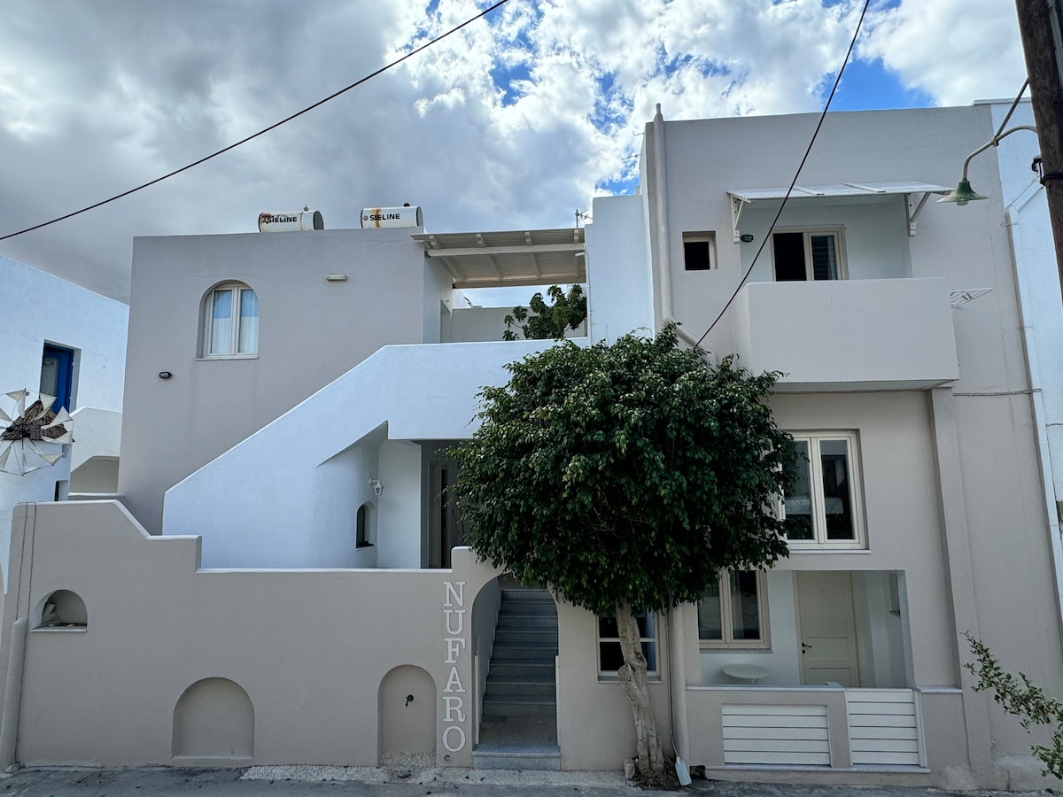 The exterior of Nufaro Studios is presented, showcasing a two-story building with a neutral color palette. Stairs lead to the entrance, flanked by a lush tree. A small sign displaying 'Nufaro' can be seen at the base, and air conditioning units are visible on a rooftop terrace.